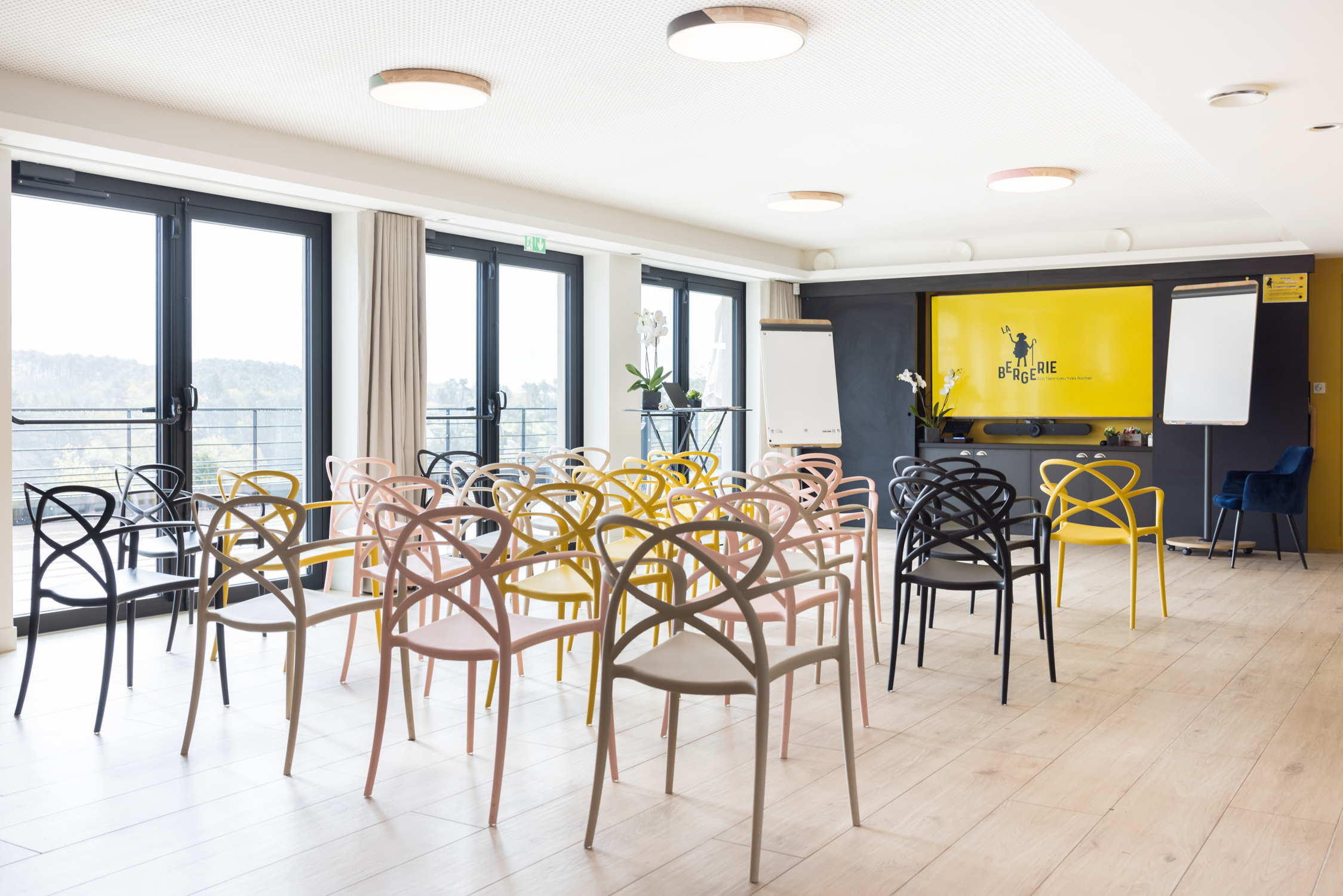 Salle de séminaire lumineuse en Bretagne avec chaises colorées, écrans de présentation et vue sur la nature à La Gacilly – Destination Yves Rocher